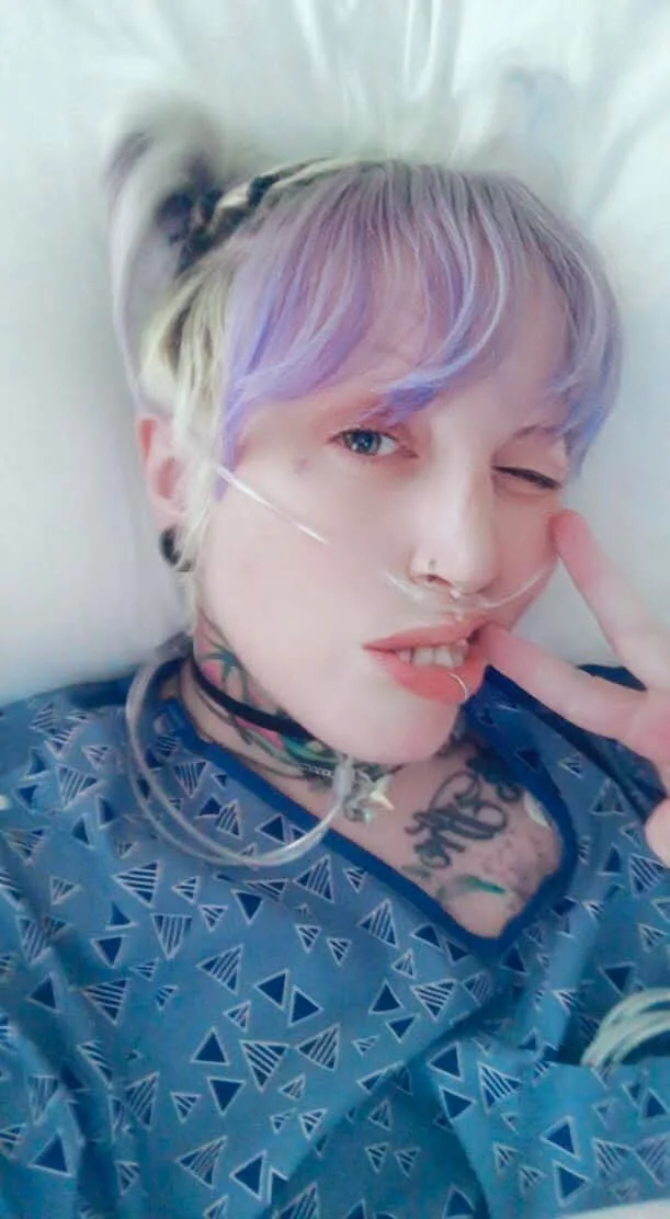A person with short, purple-tinted hair and visible tattoos lies in a hospital bed wearing a blue patterned gown. They have…