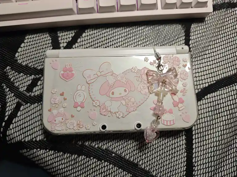 A white Nintendo 3DS console is heavily decorated with pink My Melody stickers, bows, hearts, and flowers, sitting on a…