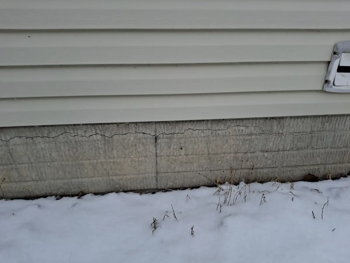 A close-up photo shows a cracked concrete foundation wall beneath beige siding, with snow and dry grass at the base. The…
