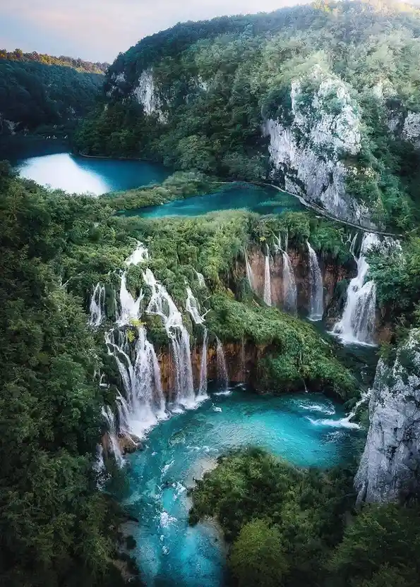 A breathtaking aerial photograph of a lush, green forest with multiple cascading waterfalls flowing into vibrant turquoise…
