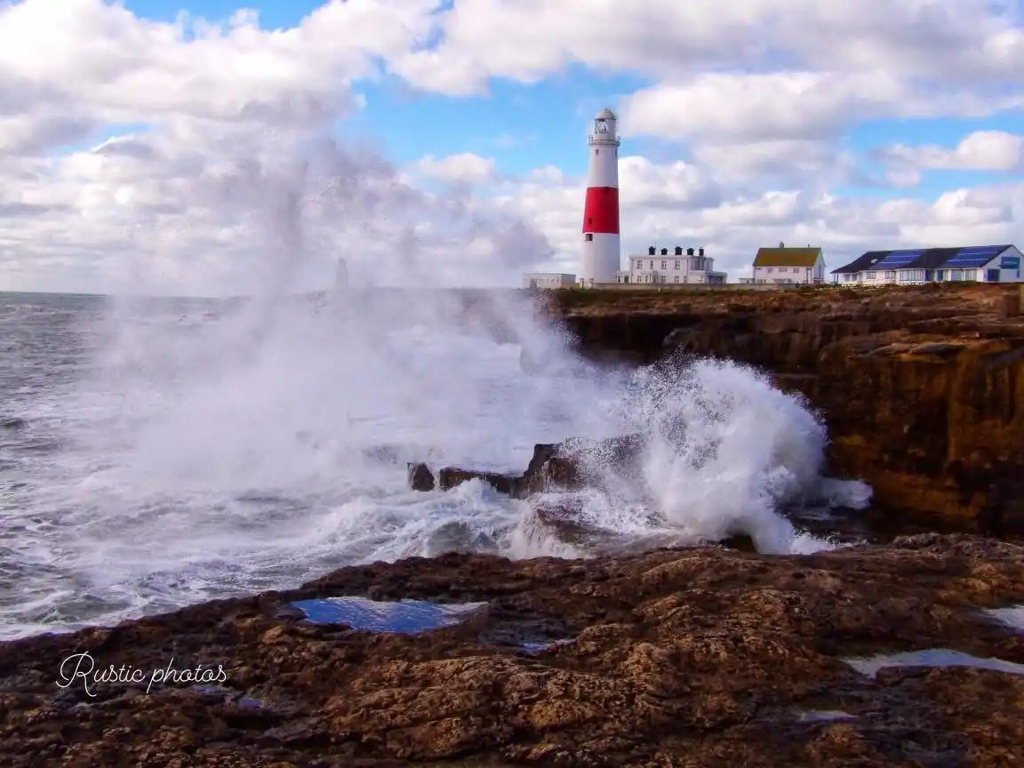 A dramatic coastal scene shows powerful waves crashing against rugged cliffs, with a red-and-white striped lighthouse…