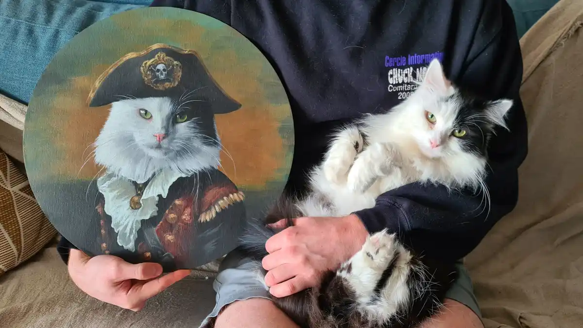 A person holds a circular portrait painting of a white and black cat dressed as a pirate, complete with a tricorn hat and…