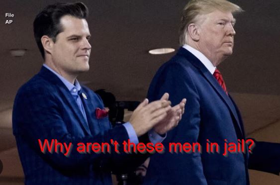 Two men in suits stand side by side; the man on the left claps while looking smugly at the man on the right, who looks away…