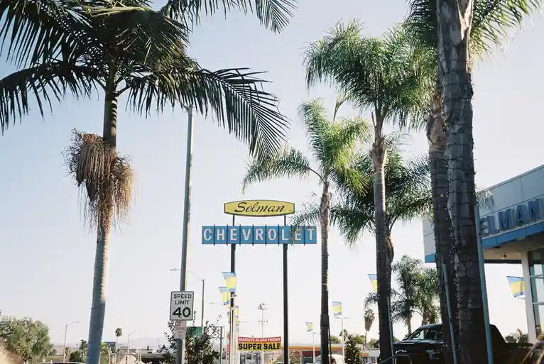 A vintage-style sign for 'Selman Chevrolet' stands prominently among tall palm trees under a clear sky, with a 'Speed Limit…