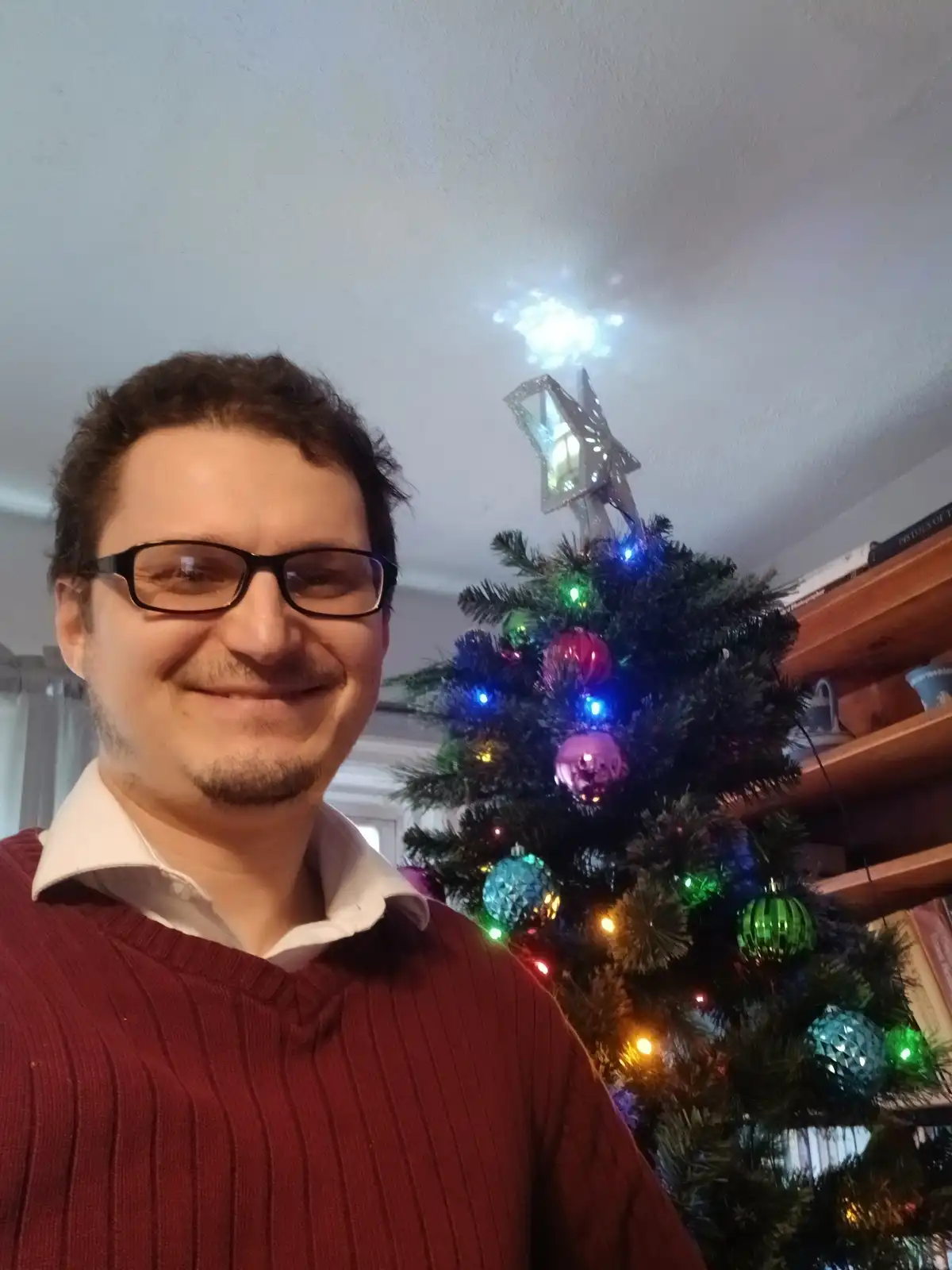 A man with glasses and a red sweater smiles at the camera while standing in front of a decorated christmas tree with…