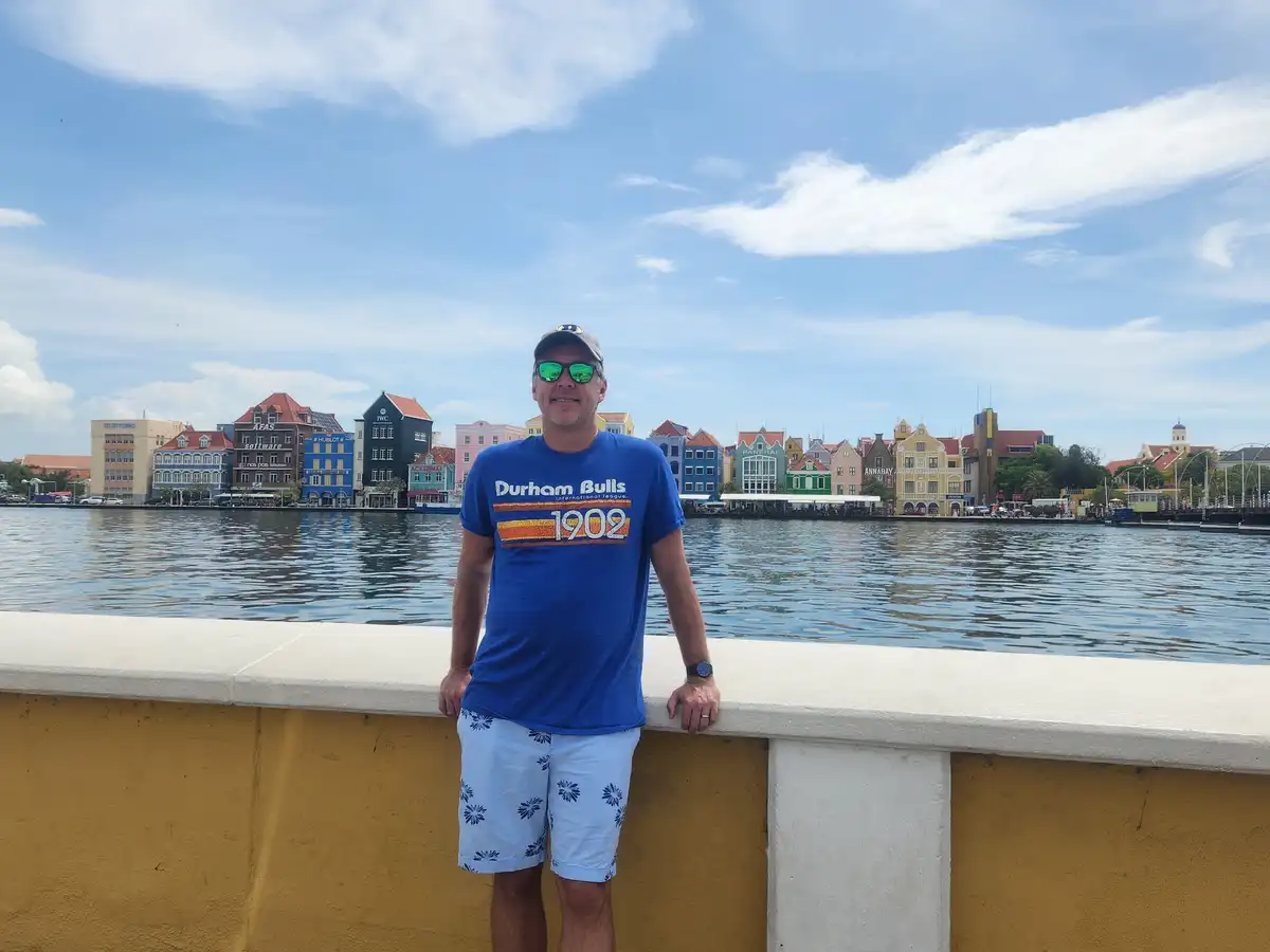 A man in a blue 'Durham Bulls 1902' t-shirt and sunglasses stands on a yellow and white waterfront wall, smiling at the…