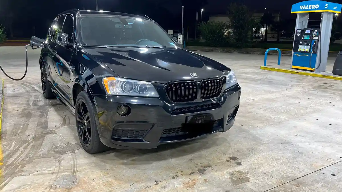 A glossy black BMW X5 SUV is parked at a Valero gas station at night, with a fuel nozzle inserted into its tank. The car's…