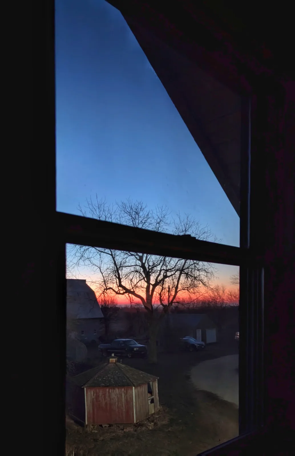 A view from inside a dark room looking out a window at a rural sunset, showing bare trees, a red shed, a barn, and parked…
