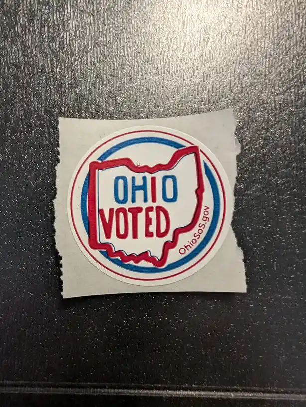 A circular Ohio state election sticker with 'OHIO VOTED' text inside a red outline of the state, set against a textured dark…