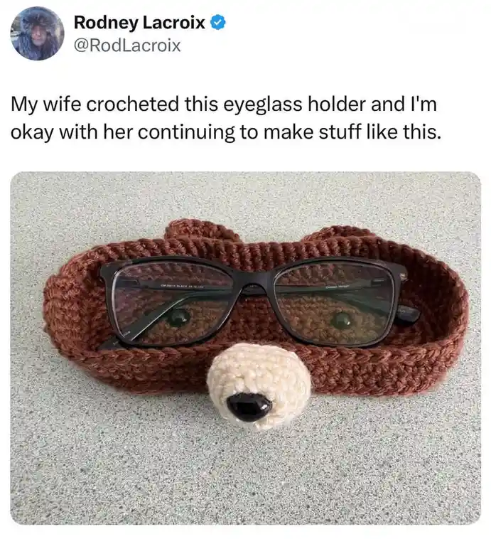 A brown crocheted eyeglass holder shaped like a bear's head, with a white snout and black button nose, holds a pair of…