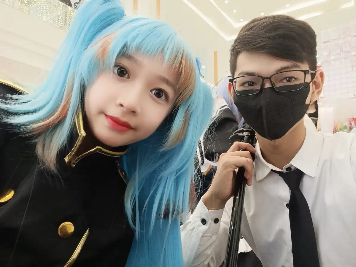 A selfie shows a young woman with bright blue hair and a black outfit posing next to a young man in a white shirt, black…