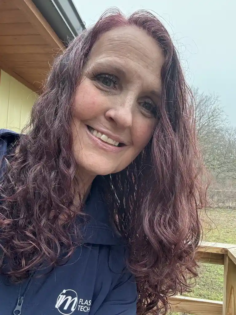 A woman with long, wavy, reddish-brown hair smiles at the camera while wearing a dark blue zip-up jacket with a white logo…