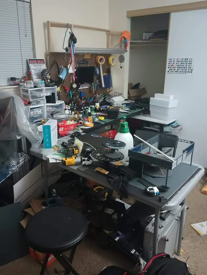 A cluttered, messy workshop or hobbyist's desk is filled with tools, parts, and supplies. A yellow drill, spools, a spray…