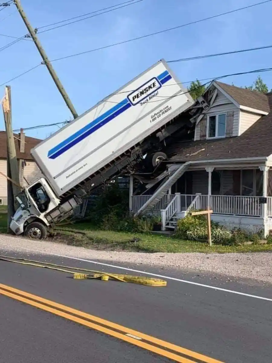 A white Penske truck rental vehicle is severely damaged, tilted at an extreme angle, with its cab crushed and the trailer…