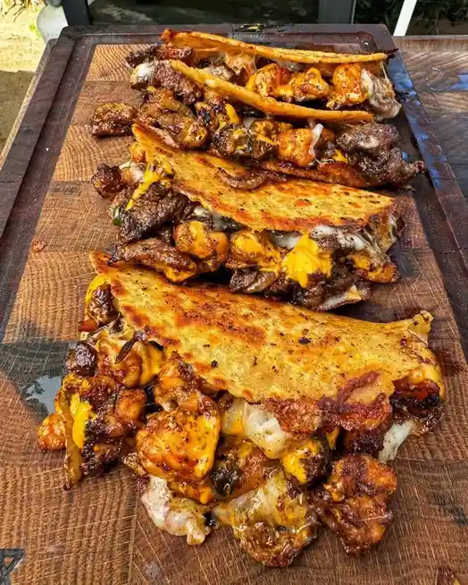 A wooden cutting board holds four large, open-faced tacos filled with melted cheese, grilled meat, and saucy ingredients…