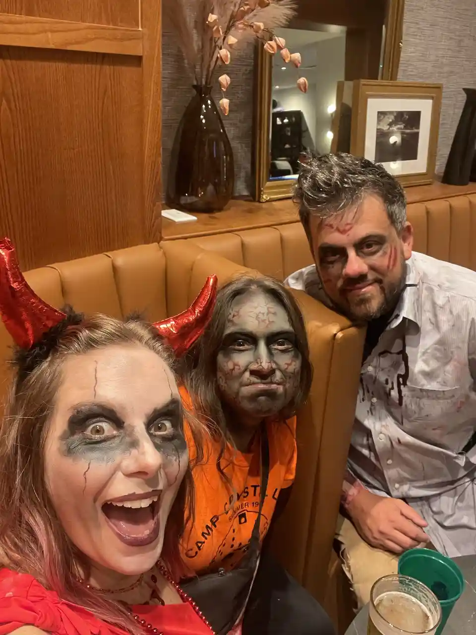 Three people in Halloween costumes sit on a brown leather couch in a bar or restaurant. The woman on the left wears red…