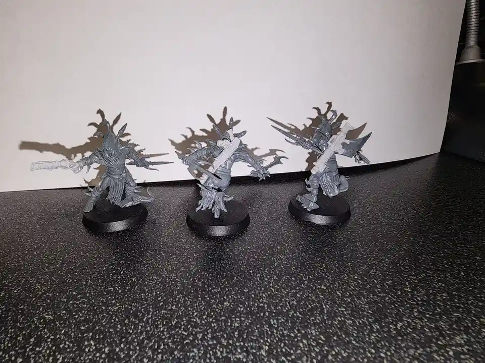 Three unpainted, grey miniature fantasy warriors stand on black circular bases on a speckled black surface, casting dramatic…