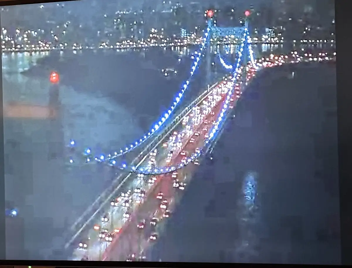 An aerial night view of a brightly lit suspension bridge with heavy traffic, showing red taillights and white headlights of…