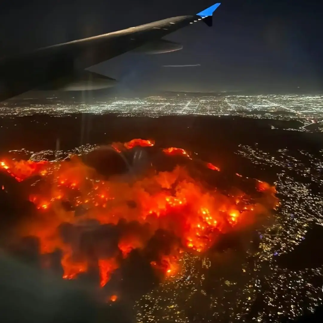 An aerial view from an airplane window shows a massive, glowing orange wildfire spreading across a dark landscape at night…