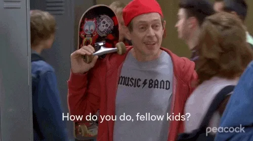 A man in a red beanie, red jacket, and gray 'MUSIC BAND' t-shirt holds a skateboard under his arm while standing in a school…