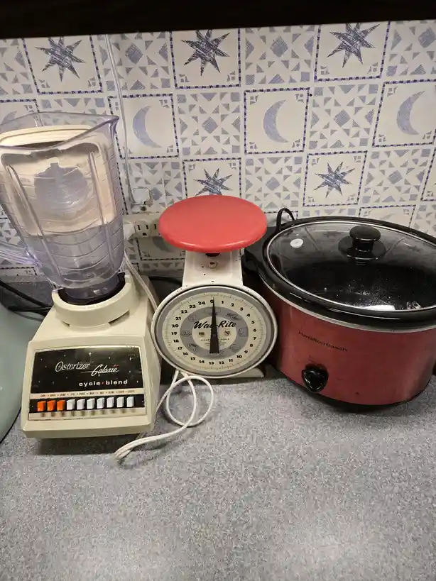 A kitchen counter displays three appliances: a beige Osterizer Galaxie blender with multiple function buttons, a vintage…