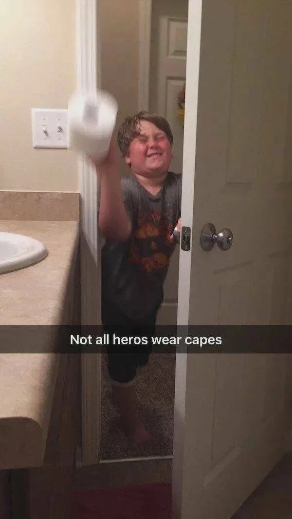 A young boy stands in a bathroom doorway, smiling broadly while holding up a large, blurry roll of toilet paper as if it…
