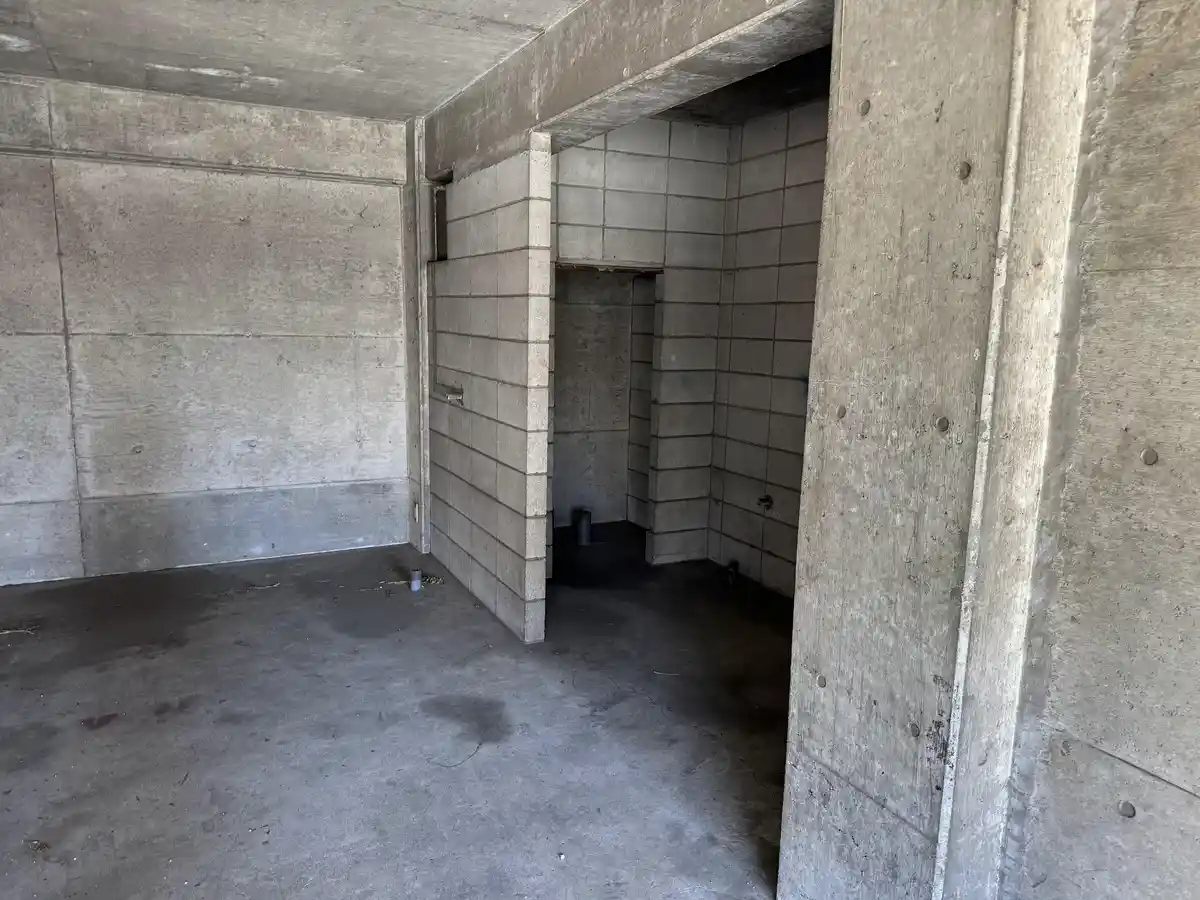 An empty, unfinished concrete room with bare walls and floor, showing exposed structural elements and a doorway leading to…