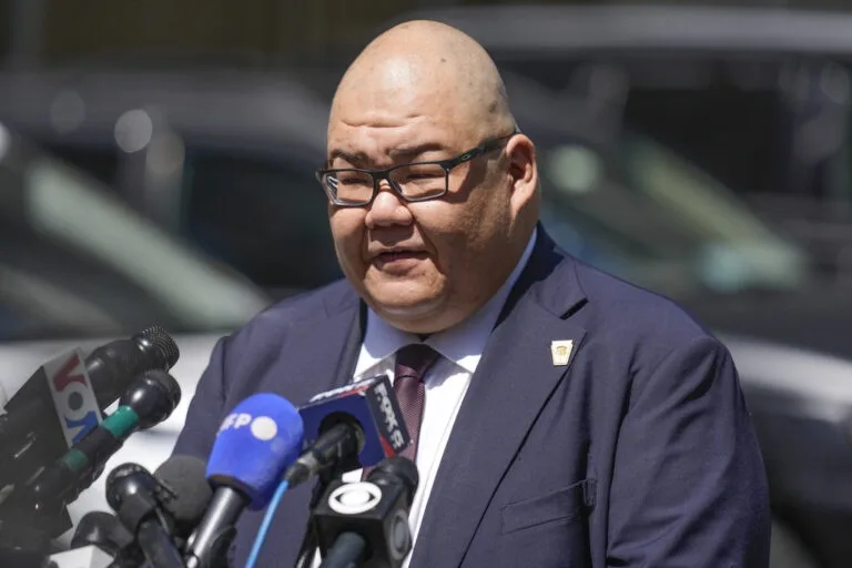 a bald man wearing glasses and a dark suit stands in front of multiple microphones from news outlets like ABC and SBS. He…