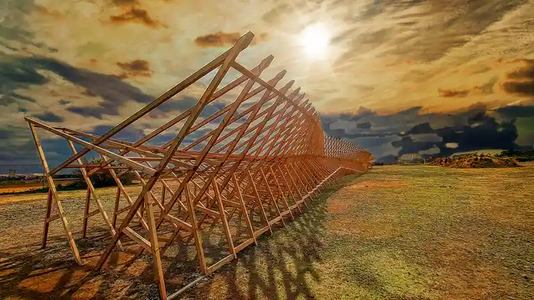 A long, zigzagging wooden fence structure stretches across a field under a dramatic, cloudy sky with the sun shining…