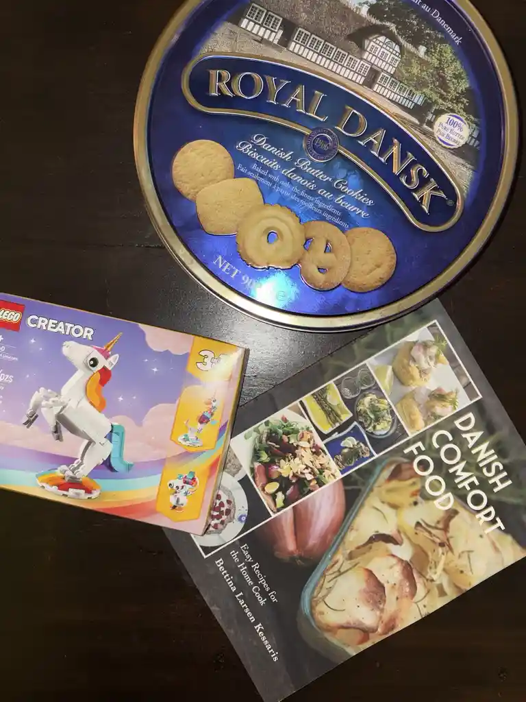 A Royal Dansk Danish Butter Cookies tin sits atop a LEGO Creator unicorn box and a 'Danish Comfort Food' cookbook, visually…