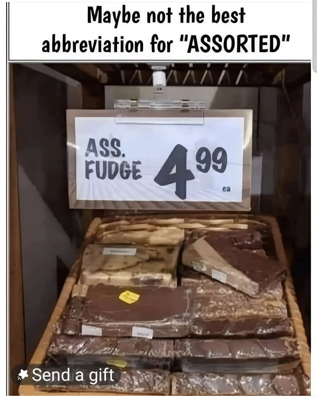 A store display of assorted fudge pieces in plastic wrapping, stacked in a wooden bin. Above them, a handwritten sign reads…