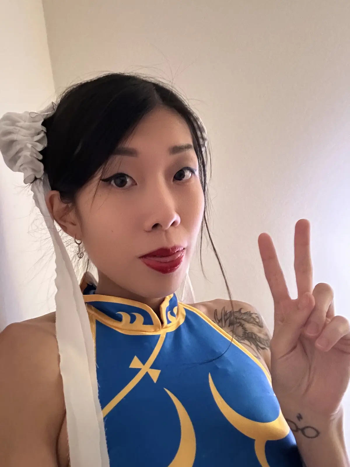 A person with dark hair tied back with a white scrunchie and red lipstick poses for a selfie, wearing a blue and yellow…