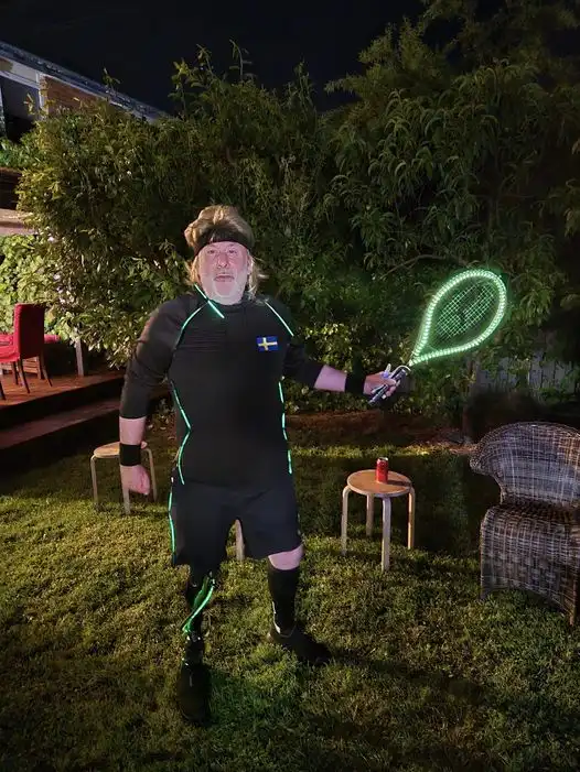 a man with a prosthetic leg and a headband stands in a backyard at night, holding a glowing green tennis racket. He wears a…