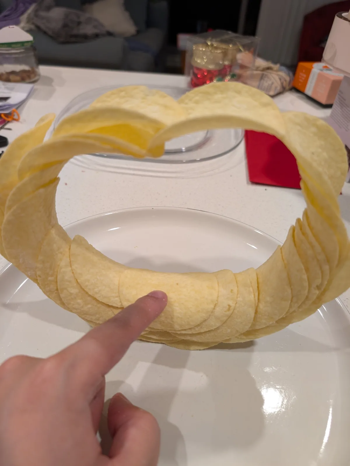 A person's finger points at a ring-shaped crown made of tortilla chips balanced on a white plate, with a blurred kitchen…