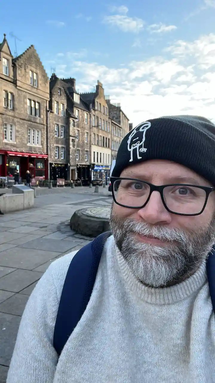 A man with glasses and a grey beard takes a selfie in a historic European square with stone buildings and a paved plaza. He…