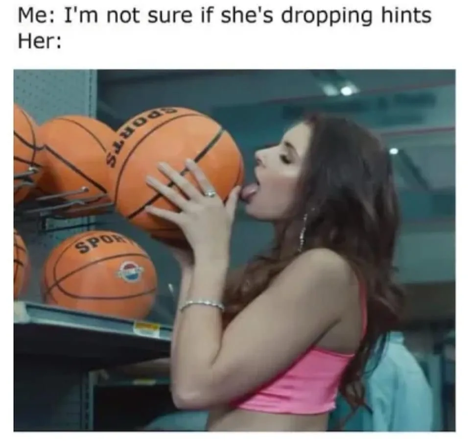 A woman in a pink top is licking an orange basketball while standing in front of a shelf full of similar basketballs. The…