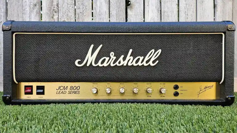 A black Marshall JCM 800 Lead Series guitar amplifier head sits on green grass with a wooden fence in the background. The…