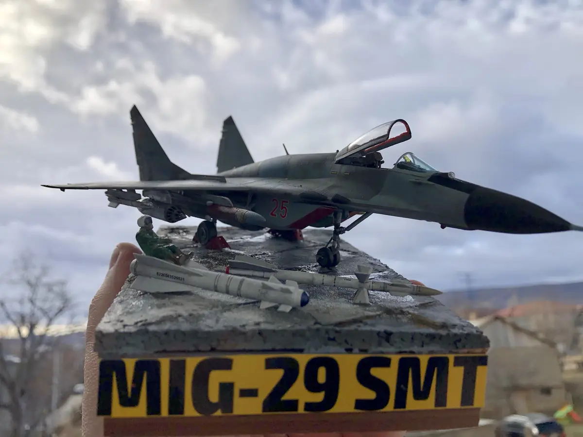 A hand holds a detailed model of a gray MiG-29 fighter jet with red number '25' on its side, mounted on a weathered concrete…