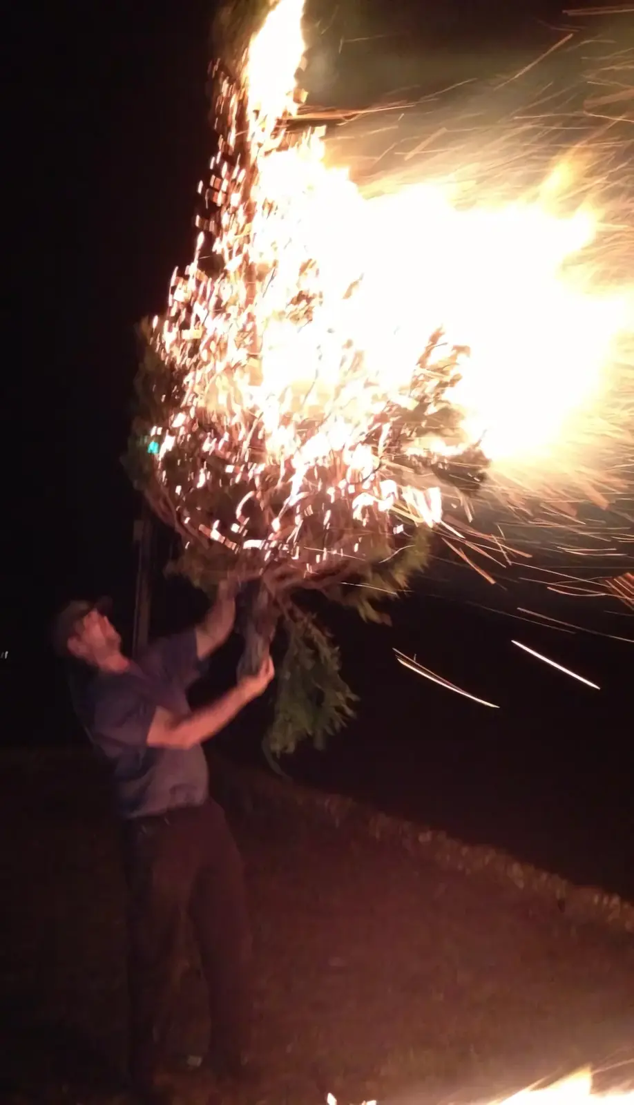A man at night holds a large evergreen tree that is engulfed in bright, fiery sparks and flames, resembling a fire dancer's…