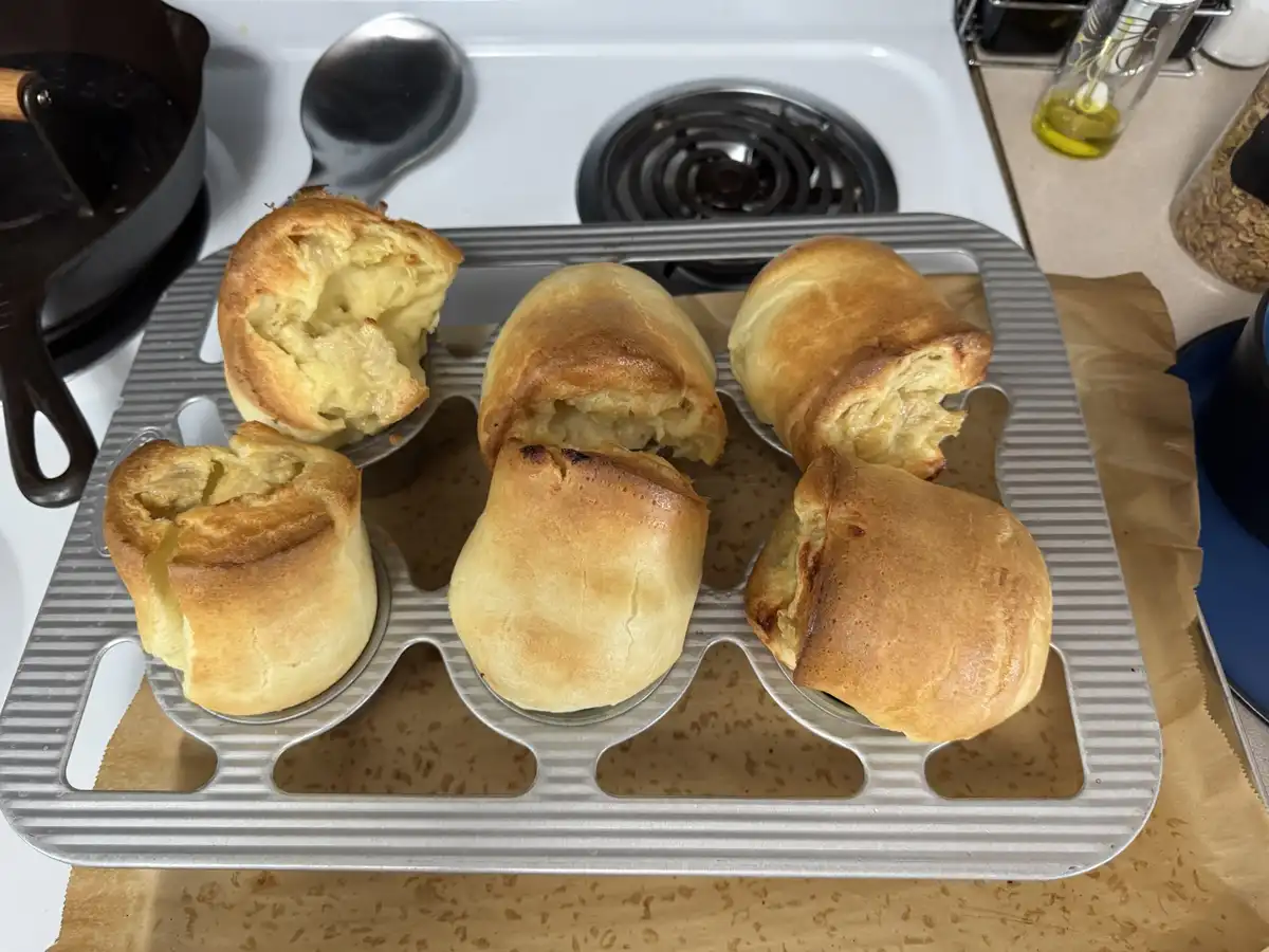 Six golden-brown popovers sit in a metal popover tin on a kitchen counter, with some visibly cracked open and risen…