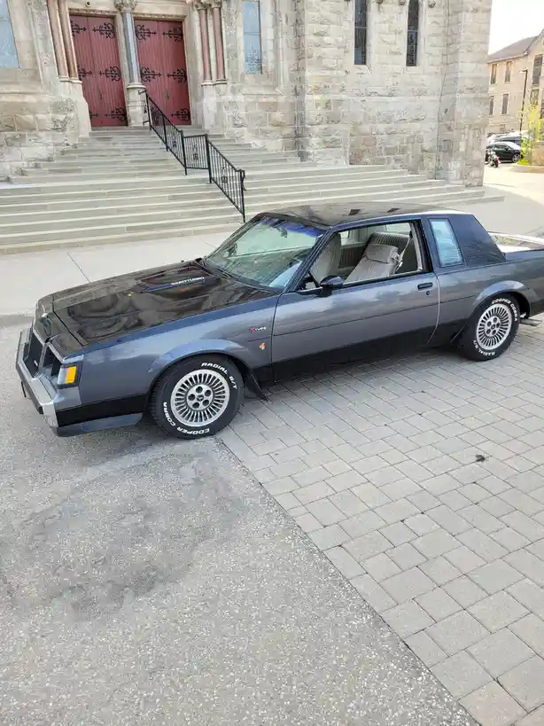 A dark gray 1980s Buick Grand National, identifiable by its '3.8L TURBO' hood decal and 'T-Type' badge, is parked on a paved…
