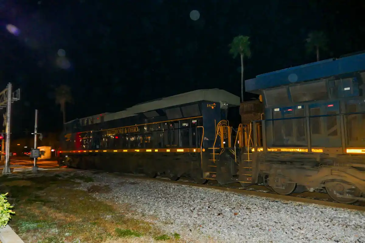 A nighttime photo of a dark blue Baltimore & Ohio train engine, number 1827, sitting on tracks next to a building with palm…