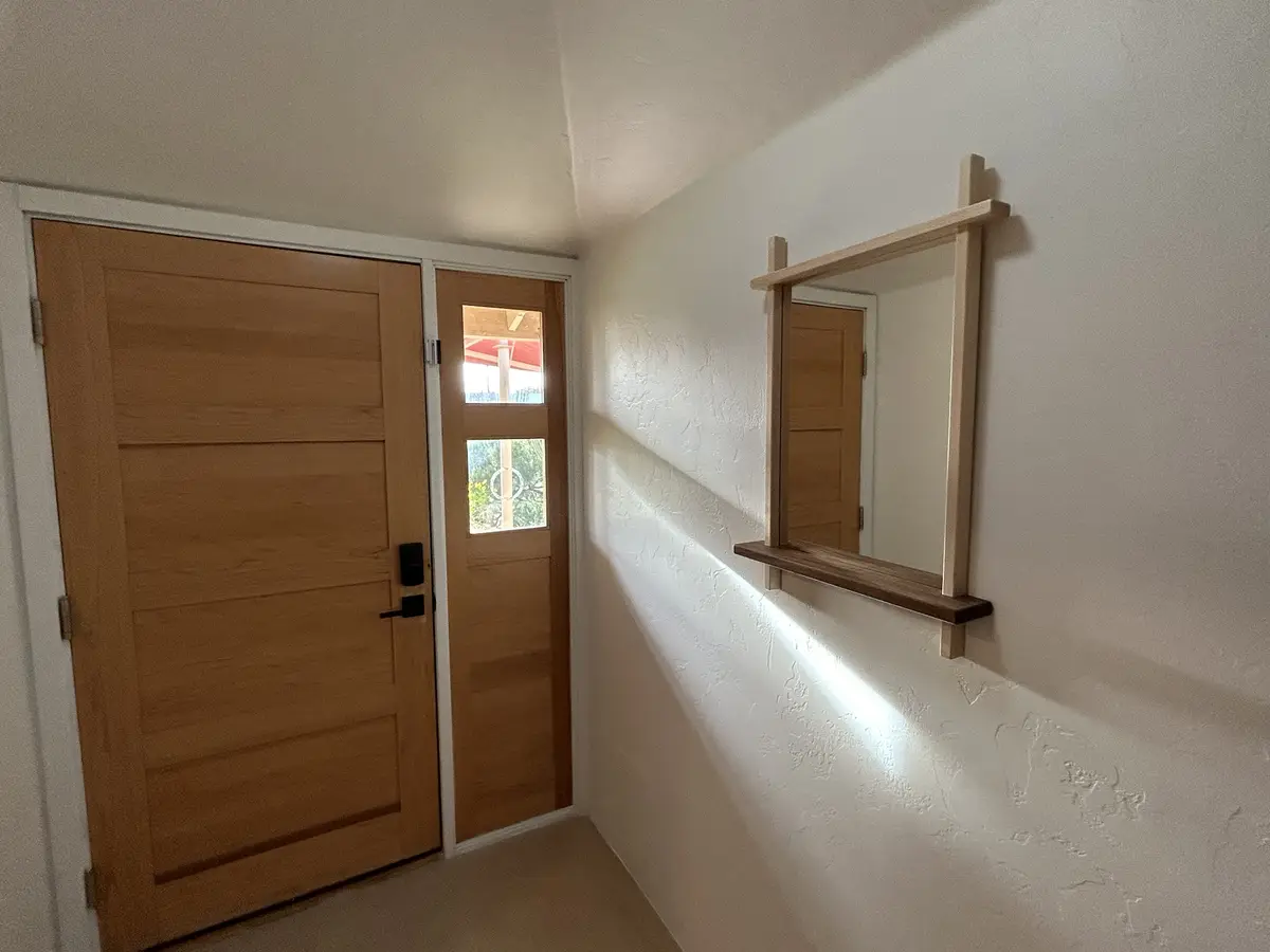 A hallway with a wooden door and a mirror mounted on the wall; the mirror reflects the door, creating a visual pun. The…