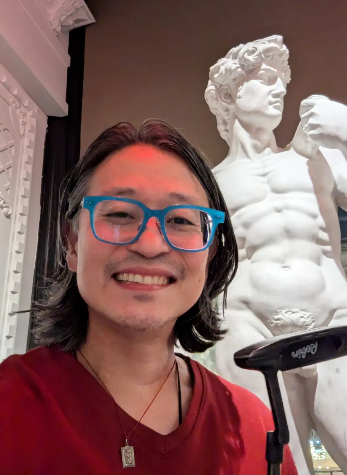 A selfie of a smiling man with blue glasses and a red shirt, standing in front of a white marble statue of David. The statue…