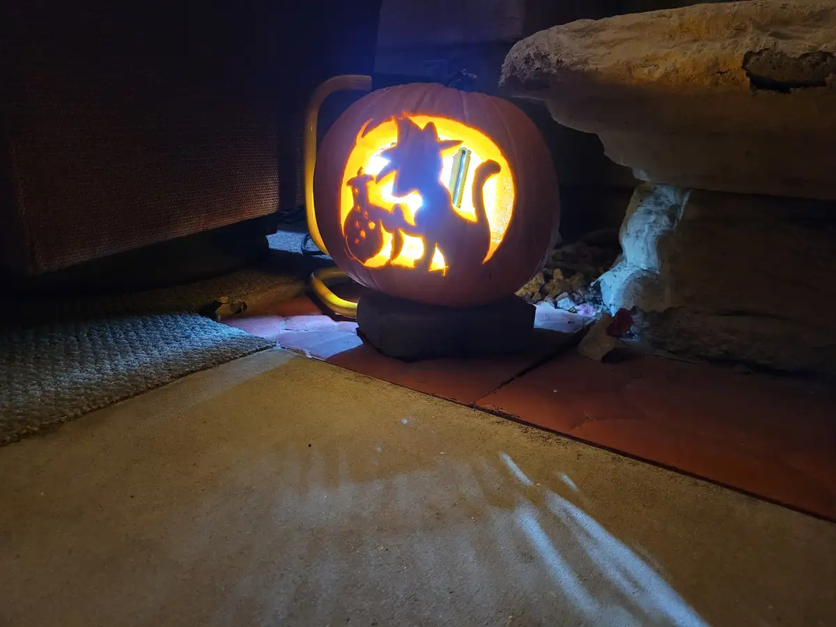 A carved Halloween pumpkin sits on a brick base, illuminated from within, casting a warm glow. The pumpkin features a…