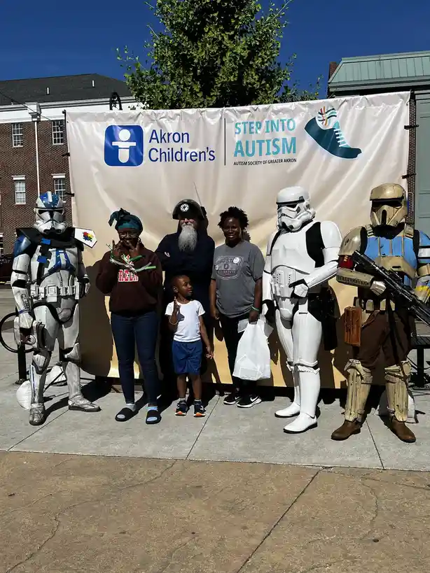 A group of people, including a child, pose for a photo in front of a banner for 'Akron Children's Step Into Autism' event…
