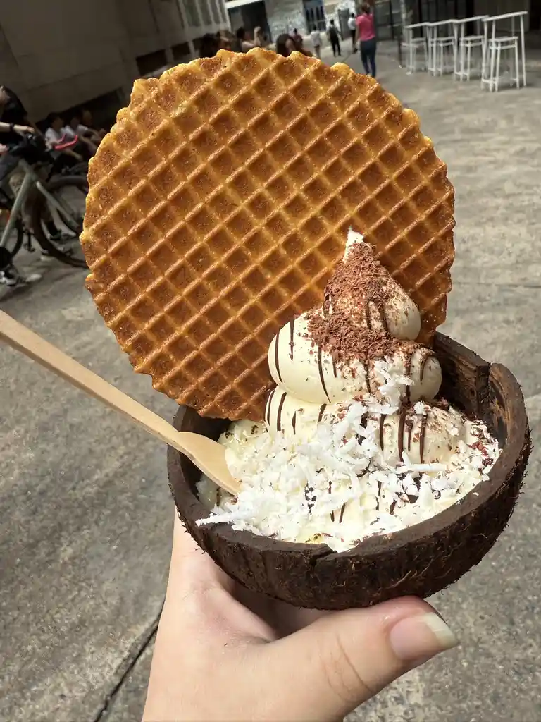 A hand holds a coconut shell filled with scoops of vanilla ice cream, topped with shredded coconut, chocolate shavings, and…