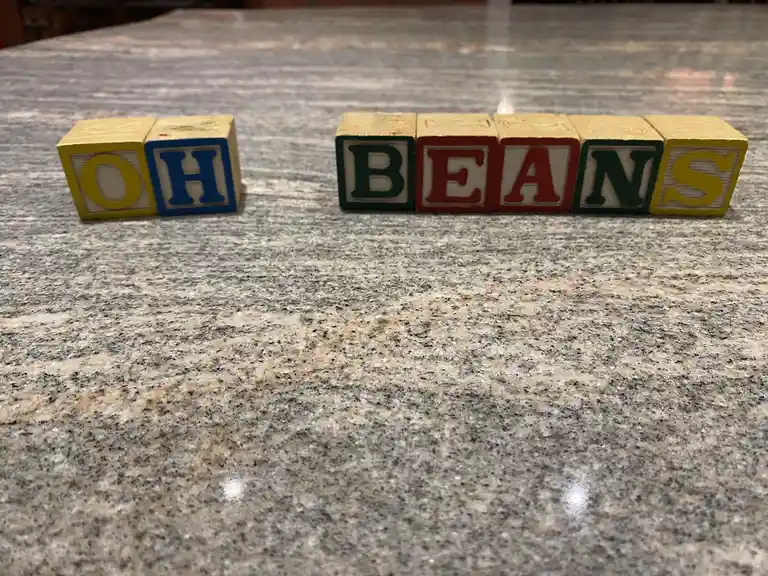 Wooden alphabet blocks arranged on a speckled granite countertop spell out 'OH' on the left and 'BEANS' on the right, with…