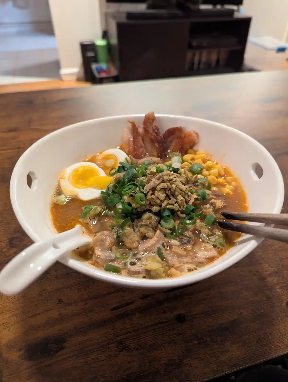 A close-up photo of a white bowl of ramen on a wooden table, filled with broth, noodles, a soft-boiled egg, crispy pork…