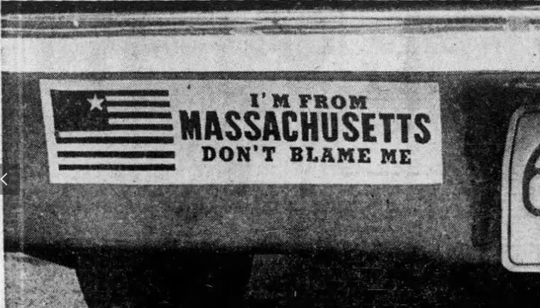 A black-and-white photo shows a bumper sticker on a car that reads 'I'M FROM MASSACHUSETTS DON'T BLAME ME' next to a…